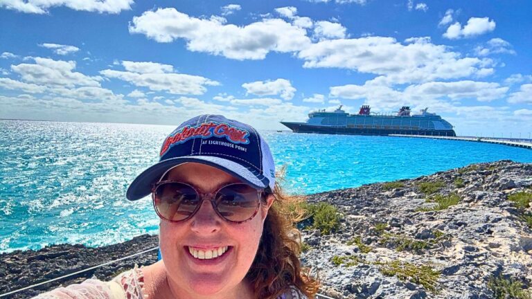 Woman in sunglasses and cap smiles with a cruise ship on clear blue water in the background.