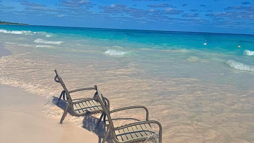 Two empty chairs on a sandy beach facing the clear blue ocean under a partly cloudy sky.