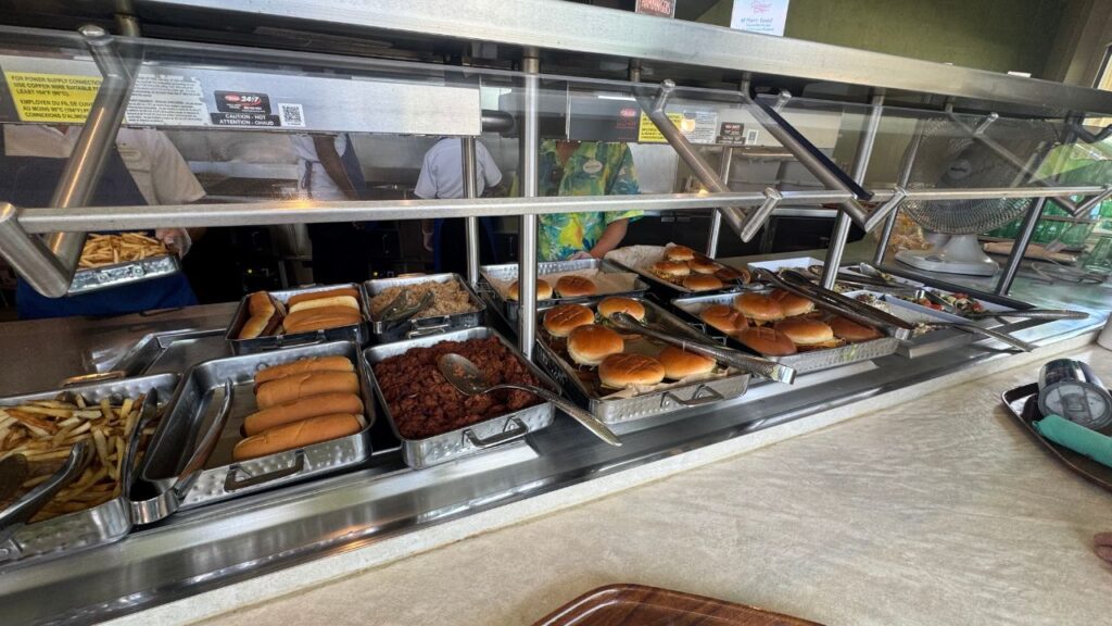 Self-serve food counter with hot dogs, burgers, fries, and various dishes in trays.