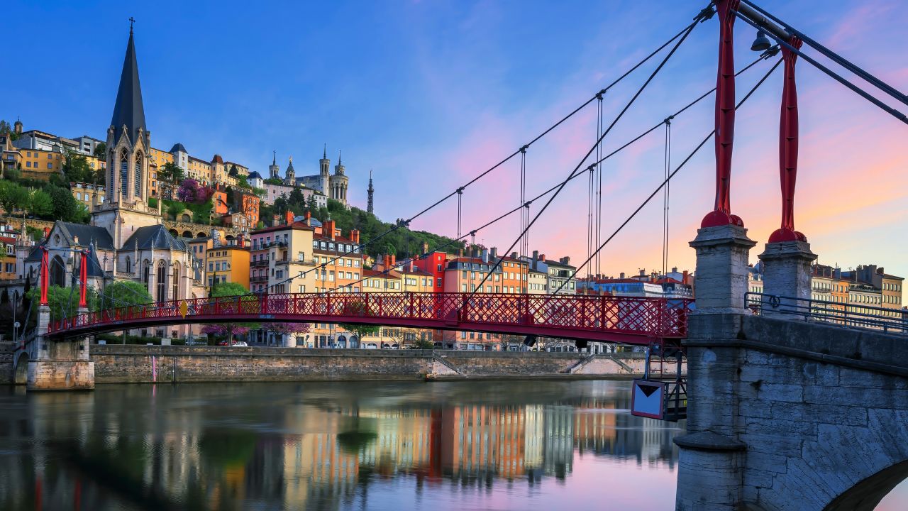 A red suspension bridge over a river, with colorful buildings and a church on a hillside in the background at sunset.