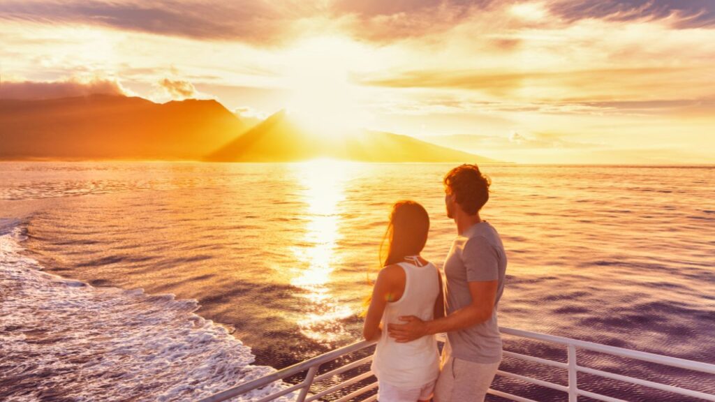 A couple stands on a boat looking at a vibrant sunset over the ocean, with mountains in the background.