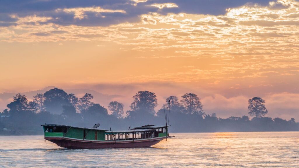 Boat on a tranquil river at sunrise, with silhouetted trees and clouds reflecting soft orange and pink hues.