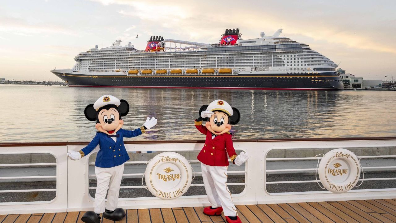 Mickey and Minnie Mouse in sailor outfits pose in front of a large cruise ship on the water at sunset.