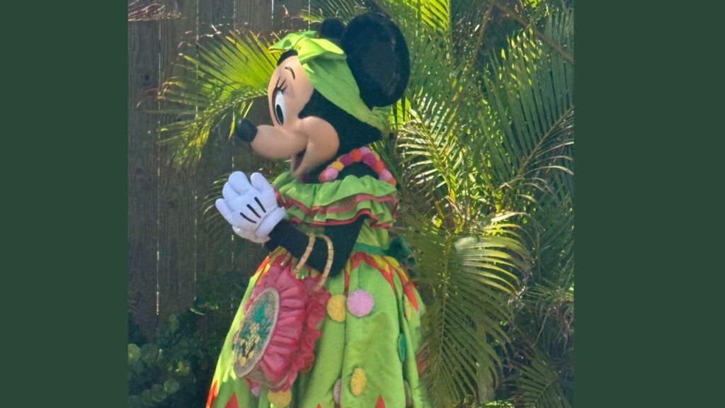 Character in a colorful green outfit with a flower theme stands in front of palm leaves outdoors.