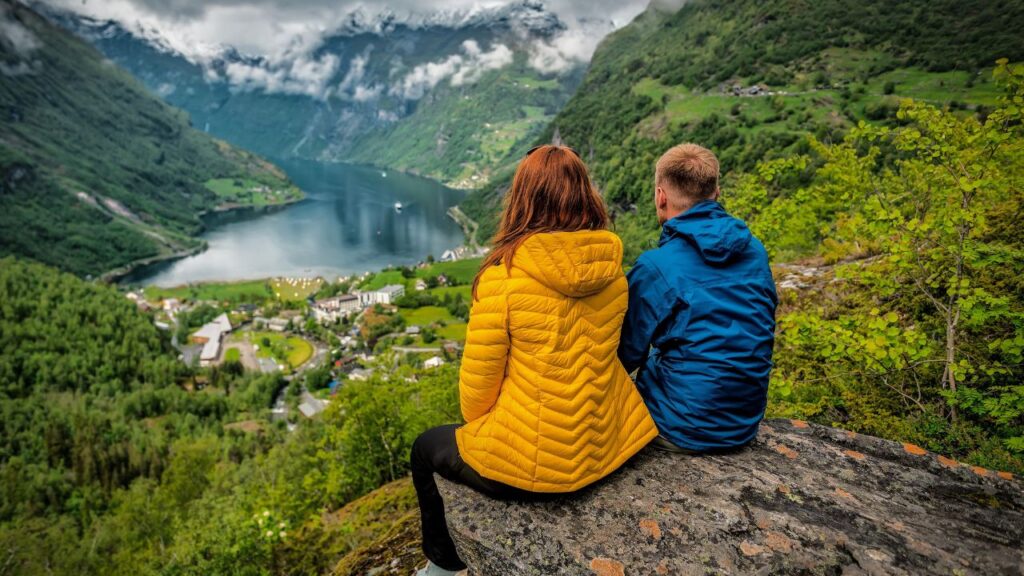 Two people in jackets sit on a rock, overlooking a scenic valley with a lake and mountains in the background.