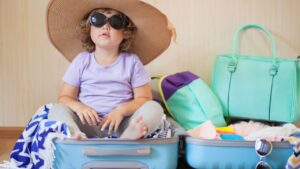 Child wearing a large sunhat and sunglasses sits in an open suitcase, surrounded by travel items.