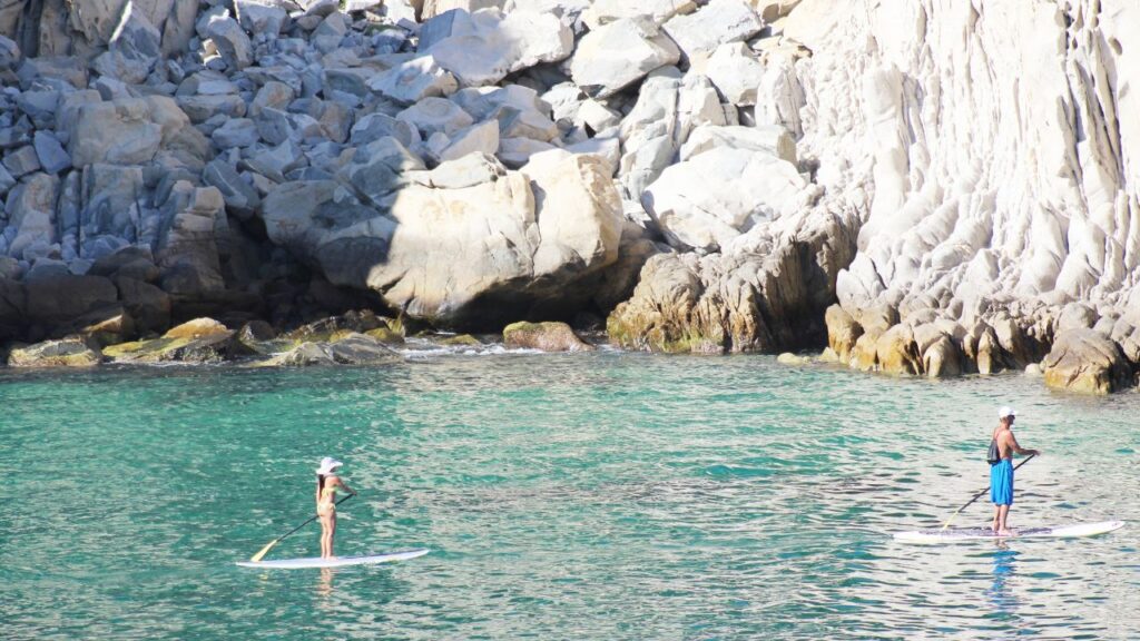 Two people paddleboarding in clear turquoise water near rocky cliffs.