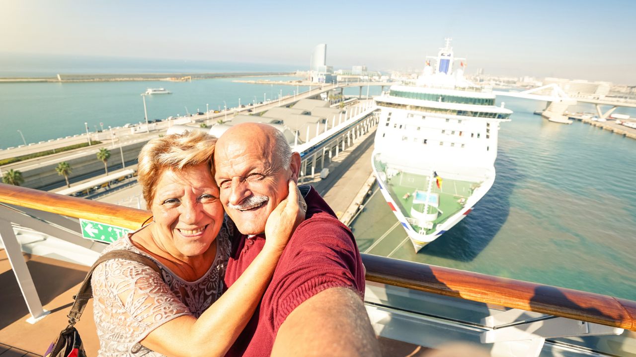 Smiling couple taking a selfie on a cruise ship deck with a port and another ship in the background.