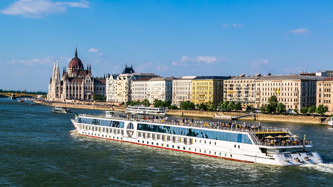 A river cruise ship sails past historic buildings and a dome on a sunny day.