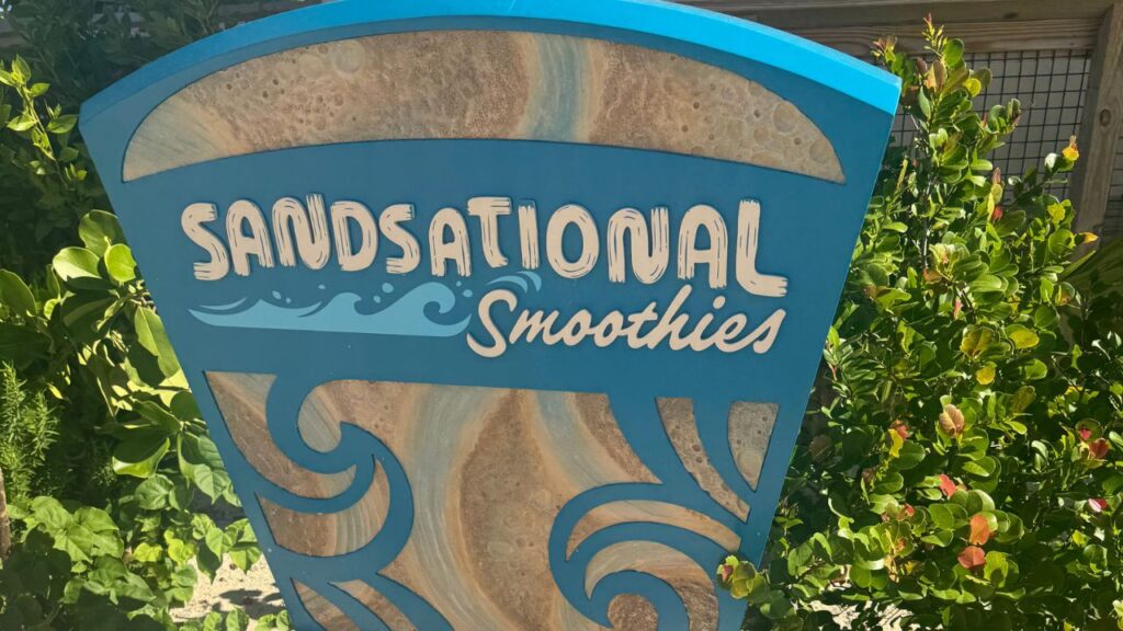 Blue sign with "Sandsational Smoothies" in white text, surrounded by lush greenery.