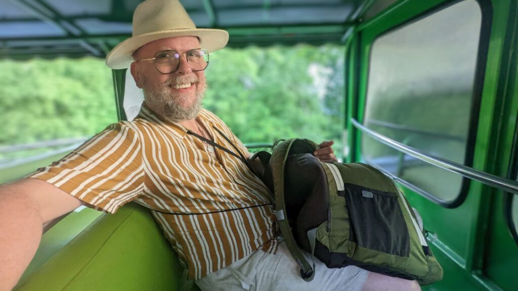 Smiling man in a striped shirt and hat sits with a backpack on a green tram.