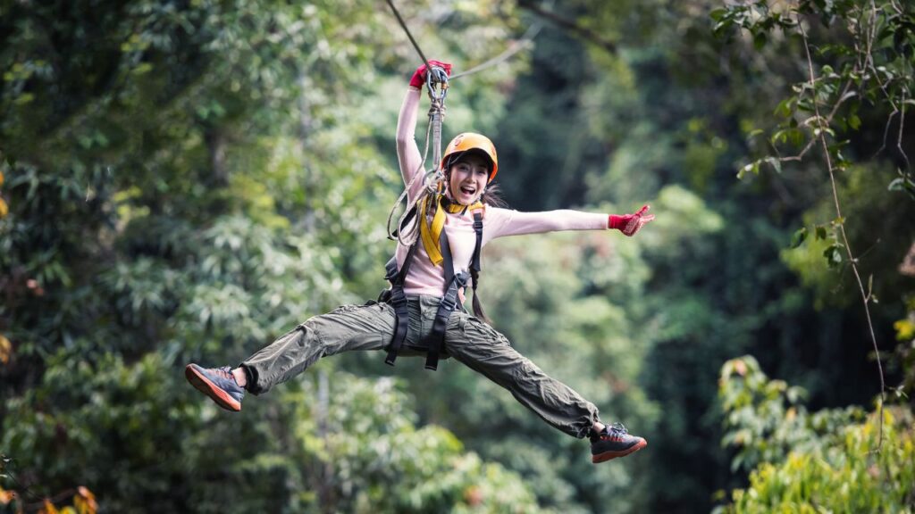 Person zip lining through a lush forest, smiling with arms and legs spread out, wearing helmet and gear.