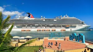 Cruise ship docked at Amber Cove, with people walking near large blue letter signs under a clear blue sky.