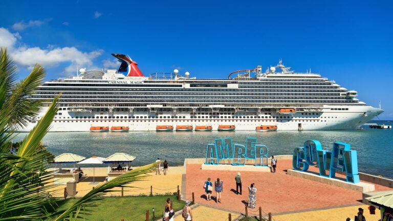 Cruise ship docked at Amber Cove, with people walking near large blue letter signs under a clear blue sky.