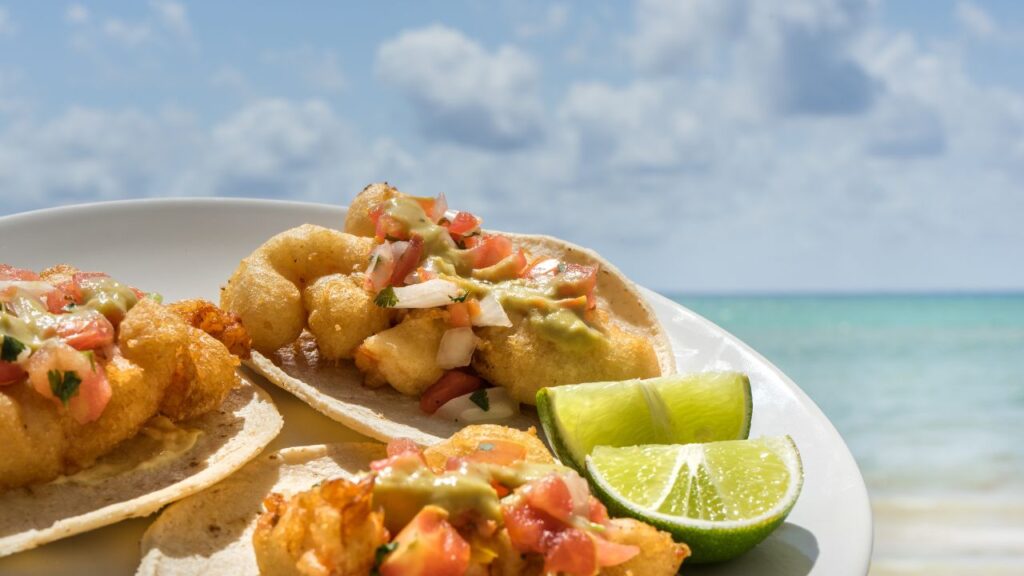 Plate of fish tacos topped with salsa and guacamole, next to lime wedges, with an ocean view in the background.