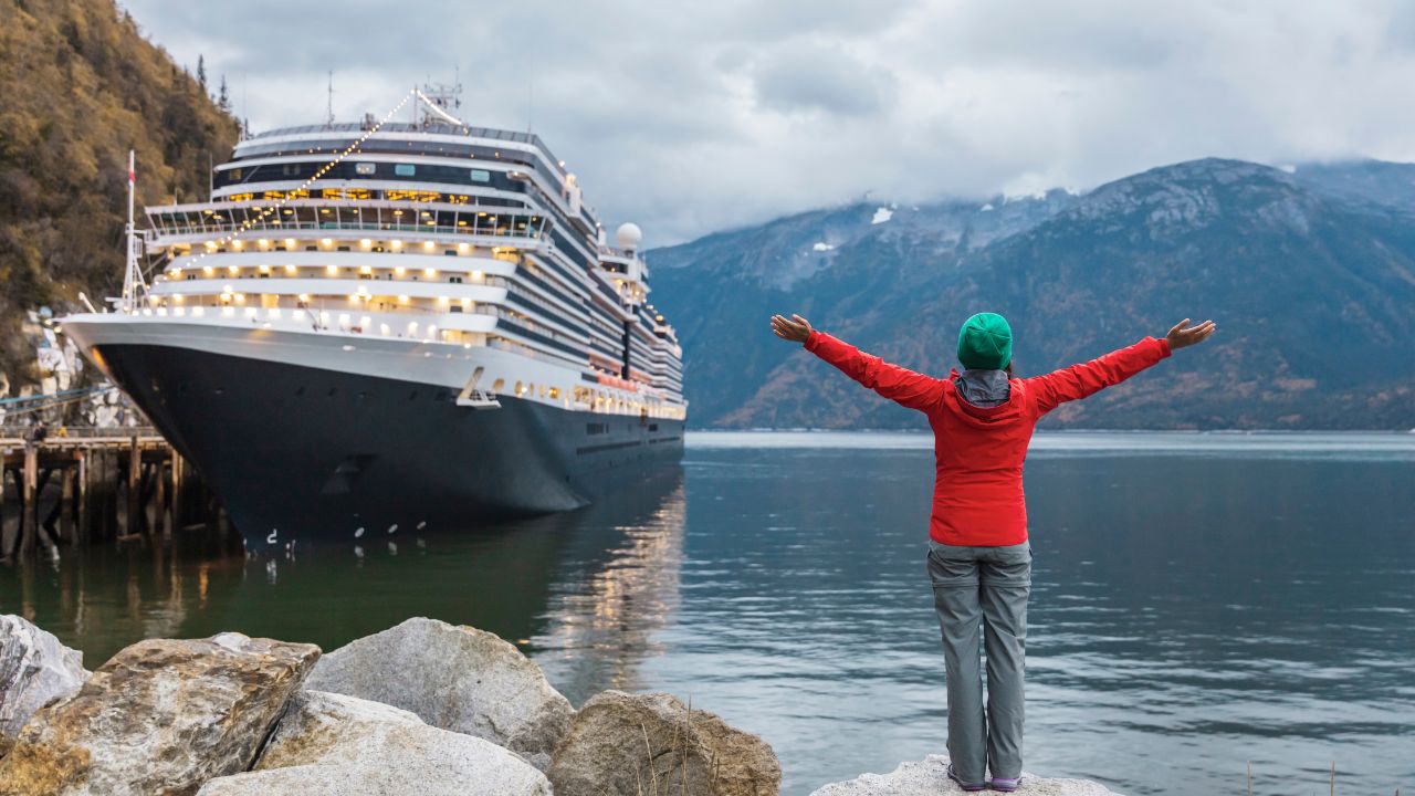 Person in red jacket with arms outstretched, facing a large cruise ship on a cloudy day in a mountainous fjord.