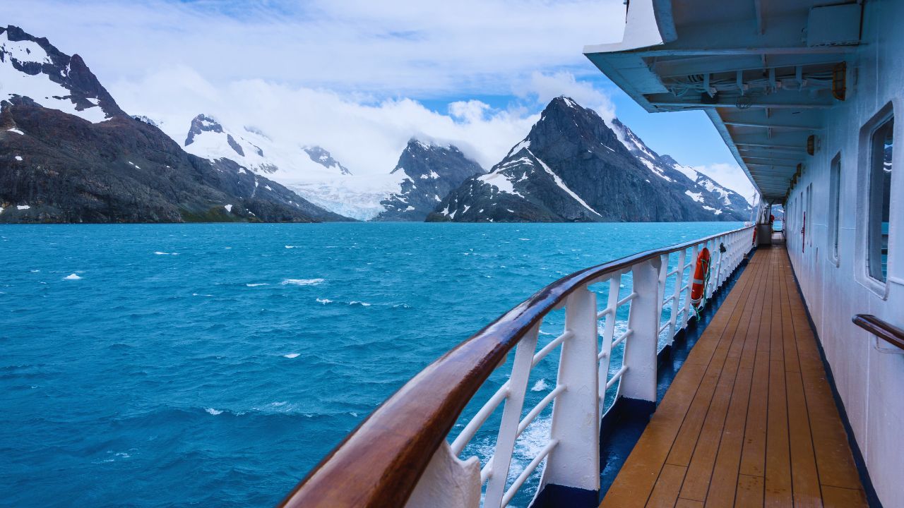 View from a ship's deck of a calm sea and snow-capped mountains under a cloudy sky.