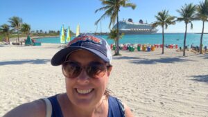 Smiling woman wearing a cap on a sunny beach with a cruise ship and palm trees in the background.