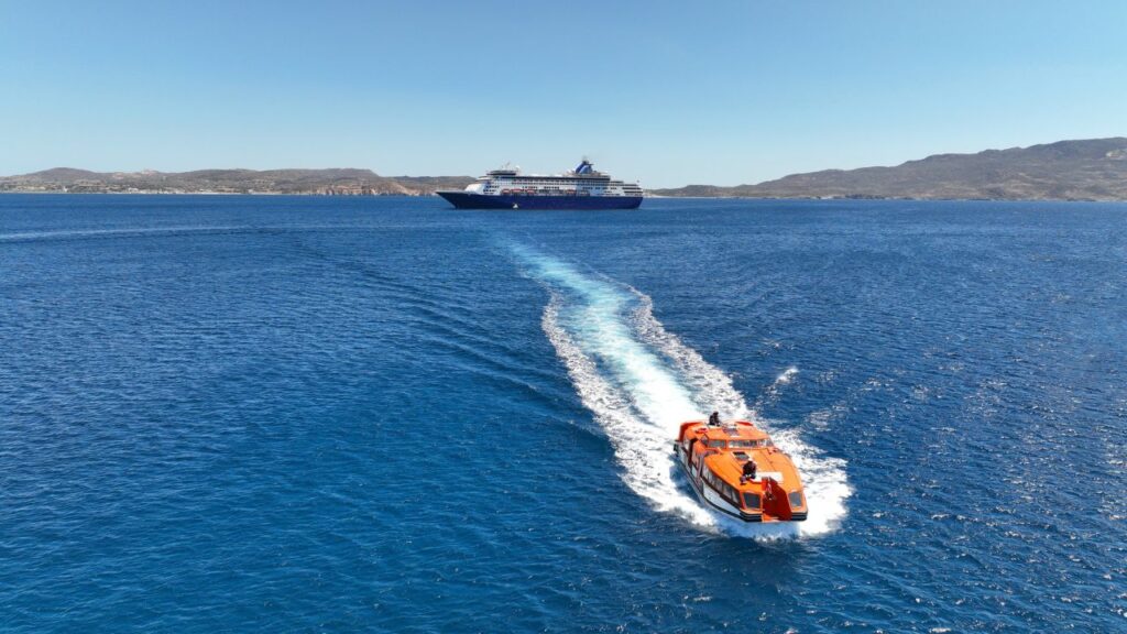 A small orange boat heads towards a cruise ship on a clear blue sea, with distant hills under a clear sky.