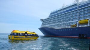 A yellow water taxi approaches a large cruise ship on a calm sea under a clear blue sky.