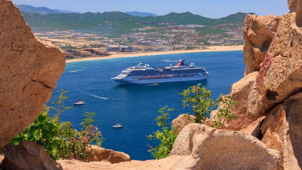 A cruise ship sails near a coastline, viewed through rocky cliffs with greenery, under a clear blue sky.