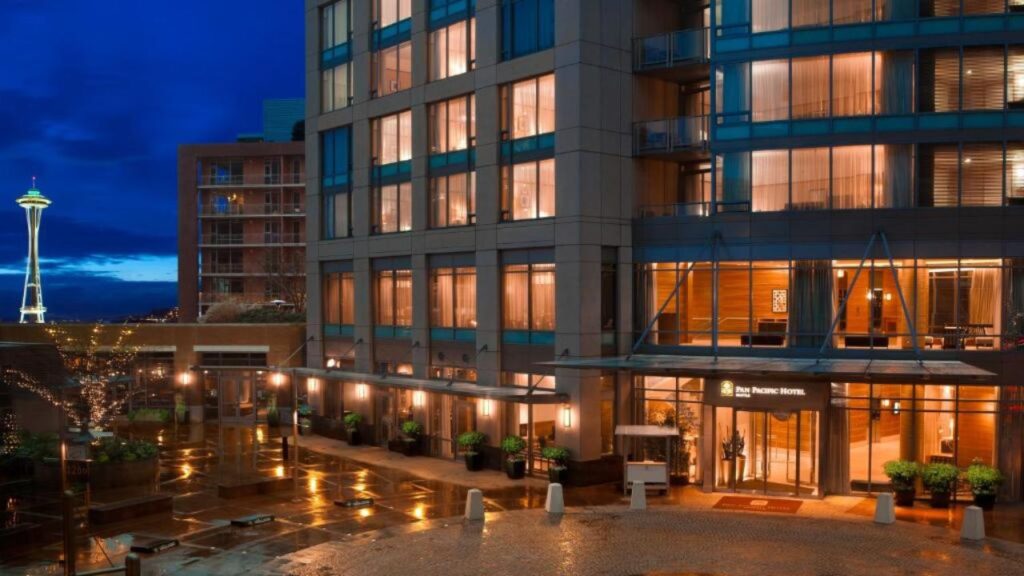 Modern hotel entrance at night with wet pavement, city lights, and Space Needle in the background.