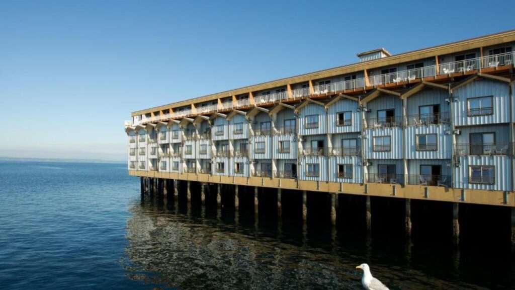 A long, light-colored building on stilts extends over calm water under a clear blue sky, with a seagull on the right.