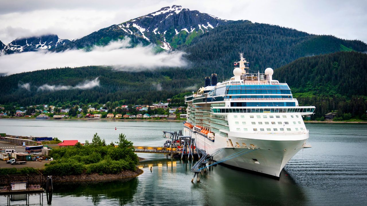 A large cruise ship docked at a port with a mountainous, forested landscape and cloudy sky in the background.