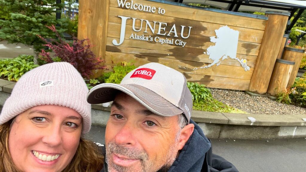 A couple smiling in front of a "Welcome to Juneau, Alaska's Capital City" sign.