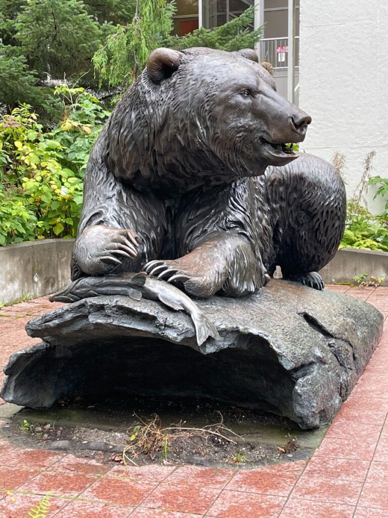 Bronze statue of a bear lying on a stone slab, surrounded by greenery.