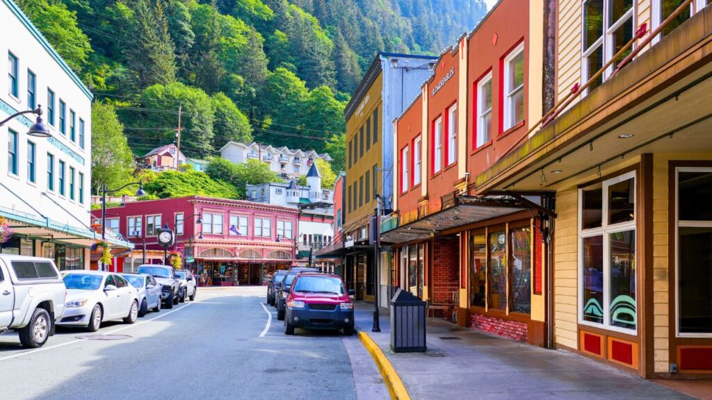 Colorful buildings line a vibrant street with parked cars, surrounded by lush green hills.