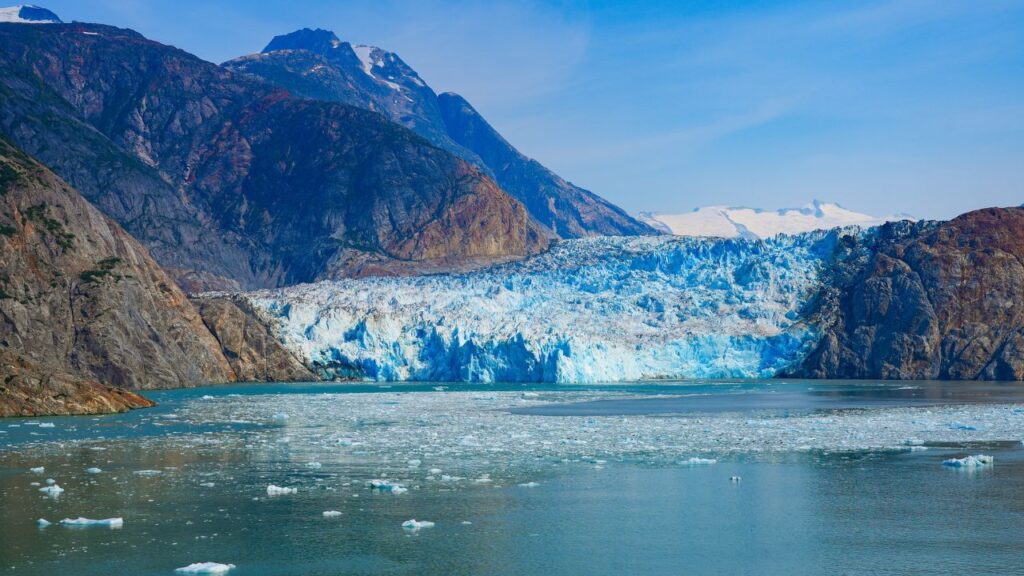 A vast blue glacier surrounded by rocky mountains, with floating ice in the clear water under a bright sky.