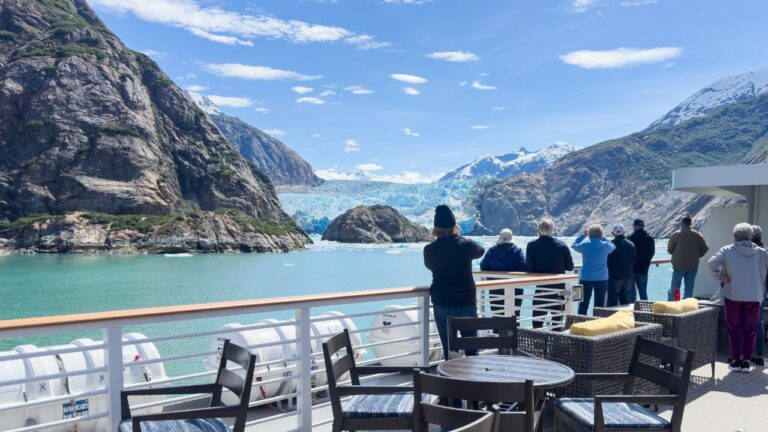 People on a ship observe glaciers and mountains under a clear blue sky.