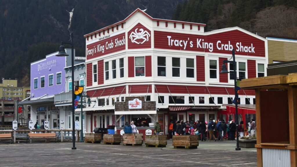 A red and white crab restaurant by a waterfront, with people queuing outside and mountains in the background.